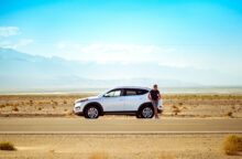 A person stands in front of a car on a stretch of road in a dessert landscape.