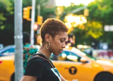 A Black woman with short curly hair looks down at her phone. There is a bright yellow taxi in the background.