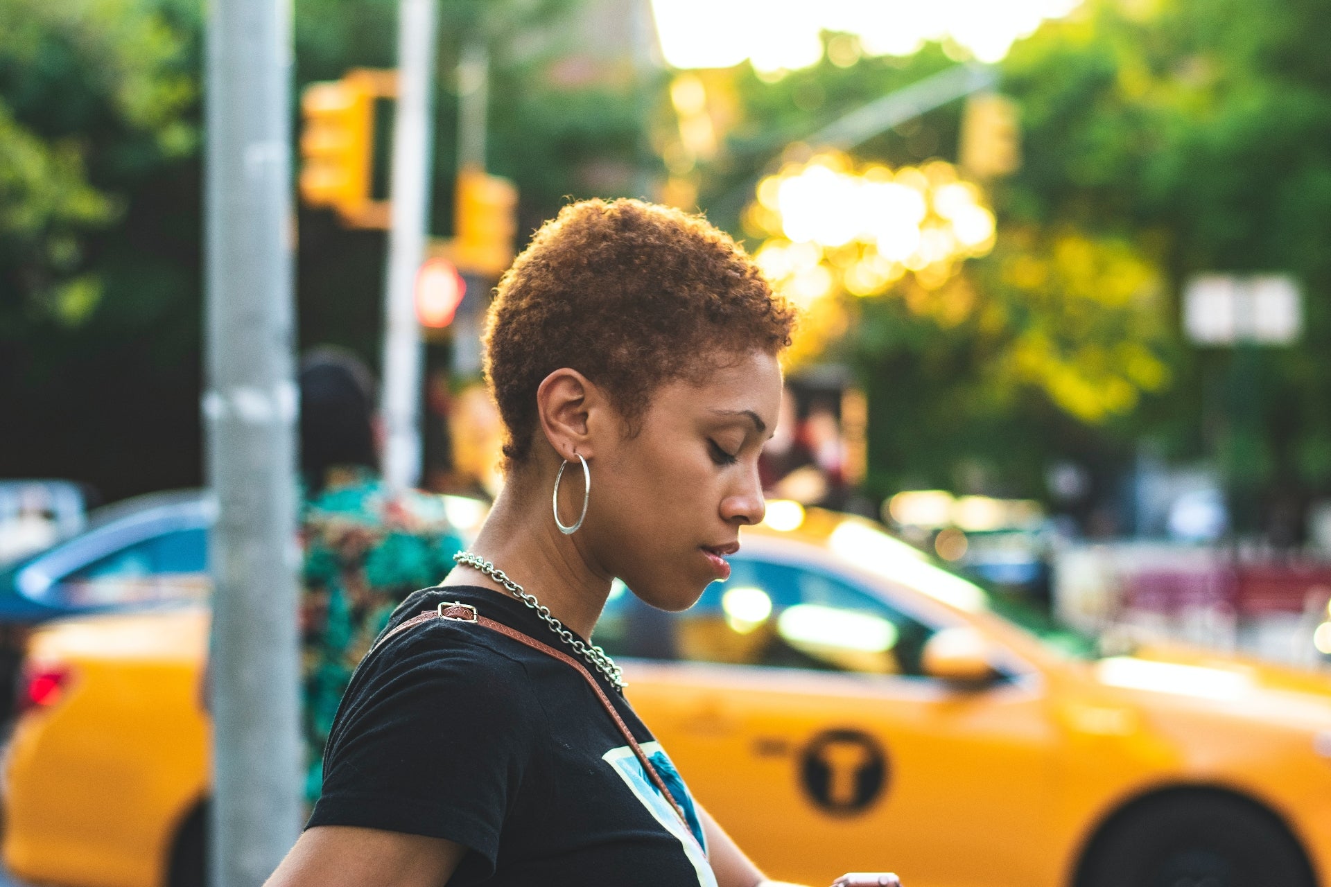 A Black woman with short curly hair looks down at her phone. There is a bright yellow taxi in the background.