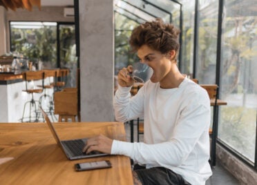 A young man sips a cup of coffee while looking at his laptop and reviewing his credit report for identity theft