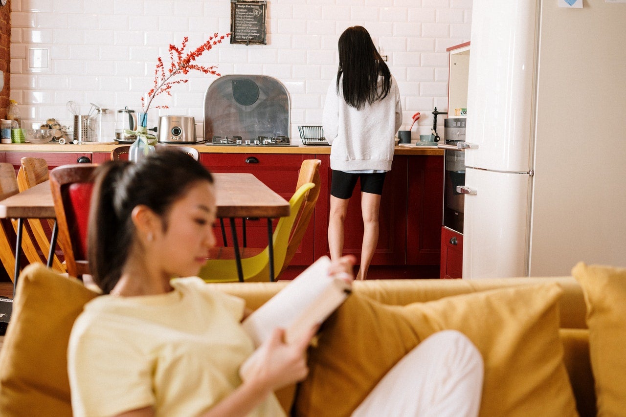 In the foreground, a roommate sits on a yellow couch reading, while in the background another roommate stands in the kitchen with their back to the camera.