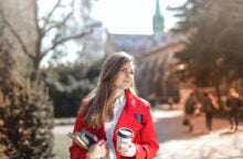 A young college girl wearing a red coat stands outside on campus, holding a stack of books and a coffee cup.