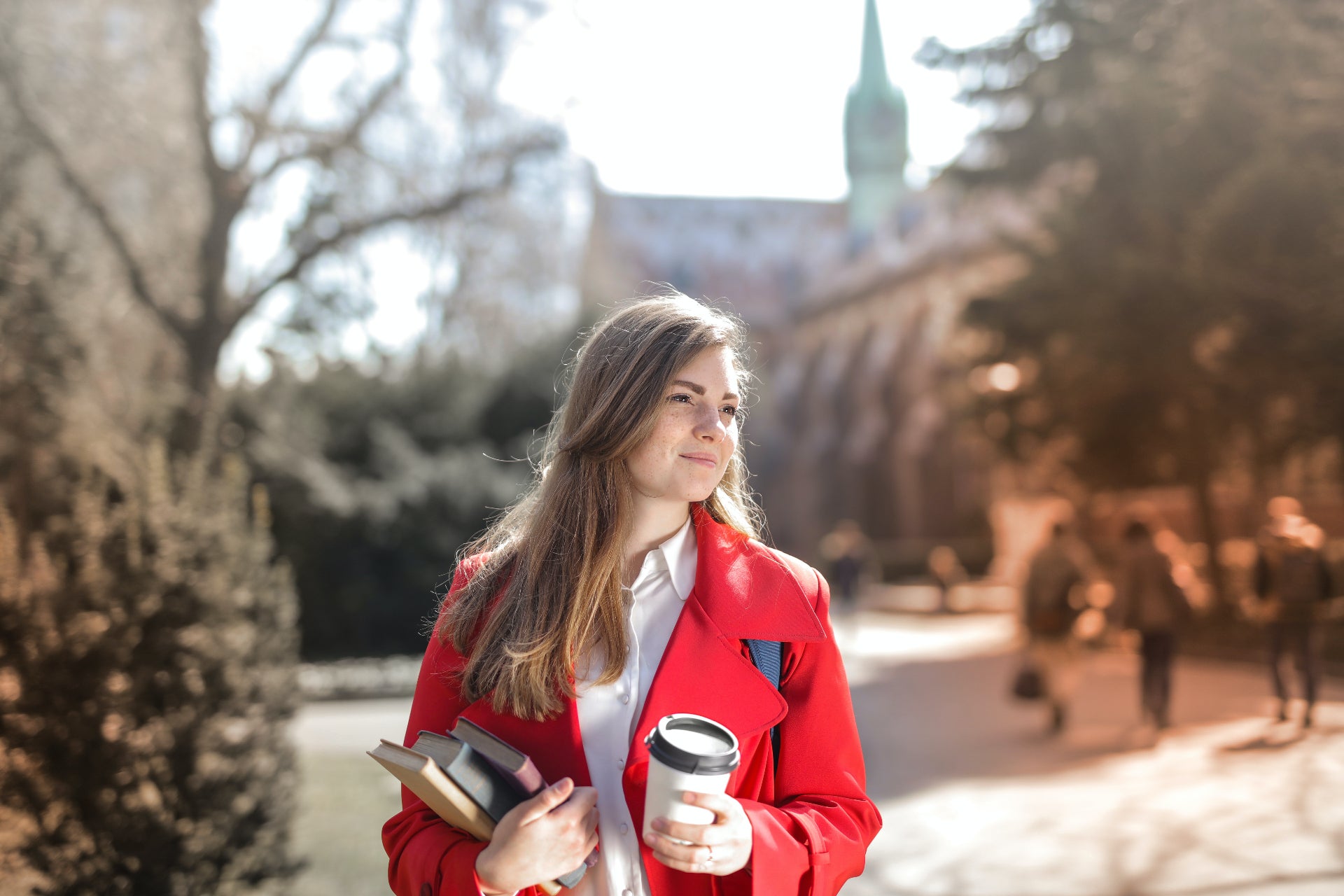 A young college girl wearing a red coat stands outside on campus, holding a stack of books and a coffee cup.