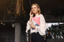 A young blond woman on a cellphone clutches a colorful notebook to her chest.