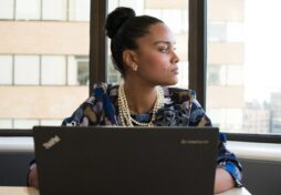 A young African American woman sits at her desk with her laptop in front of her as she looks off to the side.