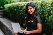 A woman sits on a bench in front of a green hedge with a laptop in her lap.