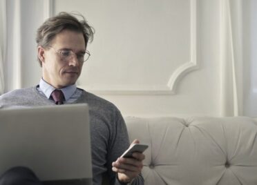 A man sits on a couch with his laptop in his lap, looking at the phone in his hand.