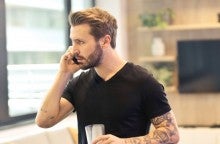 A young man with a black short sleeve shirt and arm tattoos talks on the phone with a serious look on his face