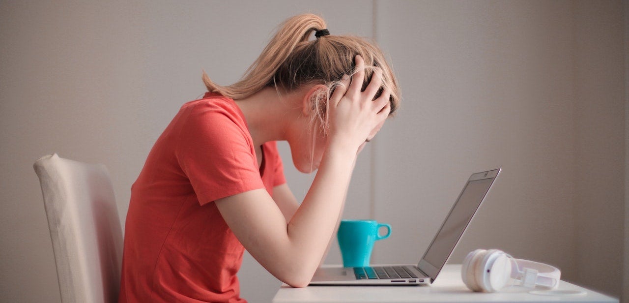 A woman in a red shirt sits in front of her laptop with her head in her hands.