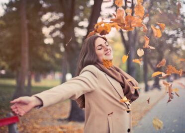 A woman wearing a brown coat and scarf smiles as she throws fall leaves around herself