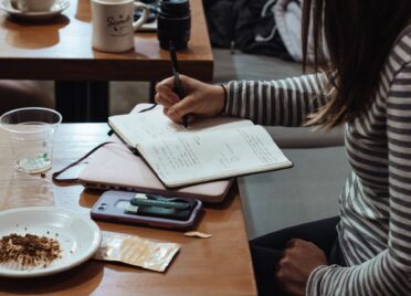 A woman wearing a longsleeved striped shirt writes in a notebook in a cafe.