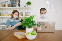 Two people in face masks sit at a kitchen table looking at their phone and laptop.