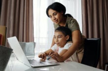 A mom discusses good money habits for kids with her daughter. The daughter is sitting at a desk and the mom is standing behind her, directing her on the laptop.