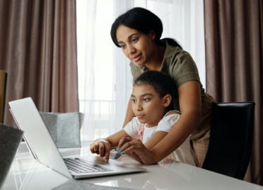A mom discusses good money habits for kids with her daughter. The daughter is sitting at a desk and the mom is standing behind her, directing her on the laptop.