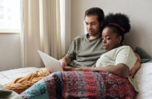 A young couple sits in bed together looking at a laptop
