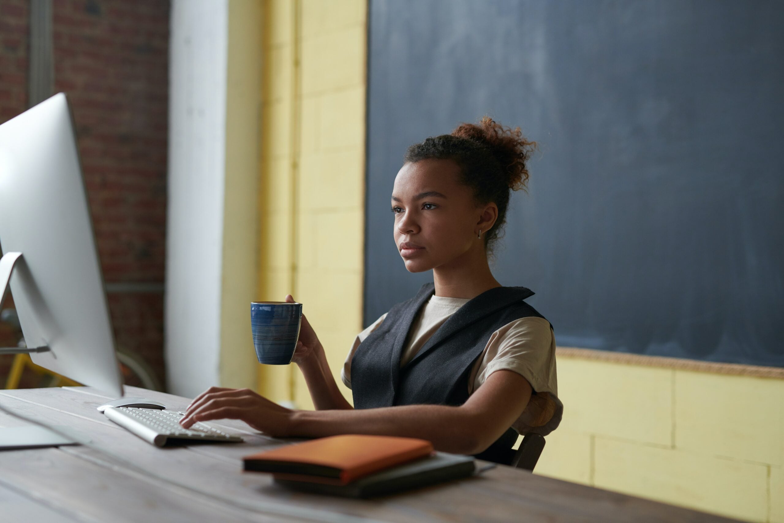 A young Black woman sits at a desktop computer and looks intently at the screen. One hand rests on the keyboard and the other holds a mug of coffee or tea. There is a yellow wall and blackboard behind her and two notebooks stacked on the desk beside her.