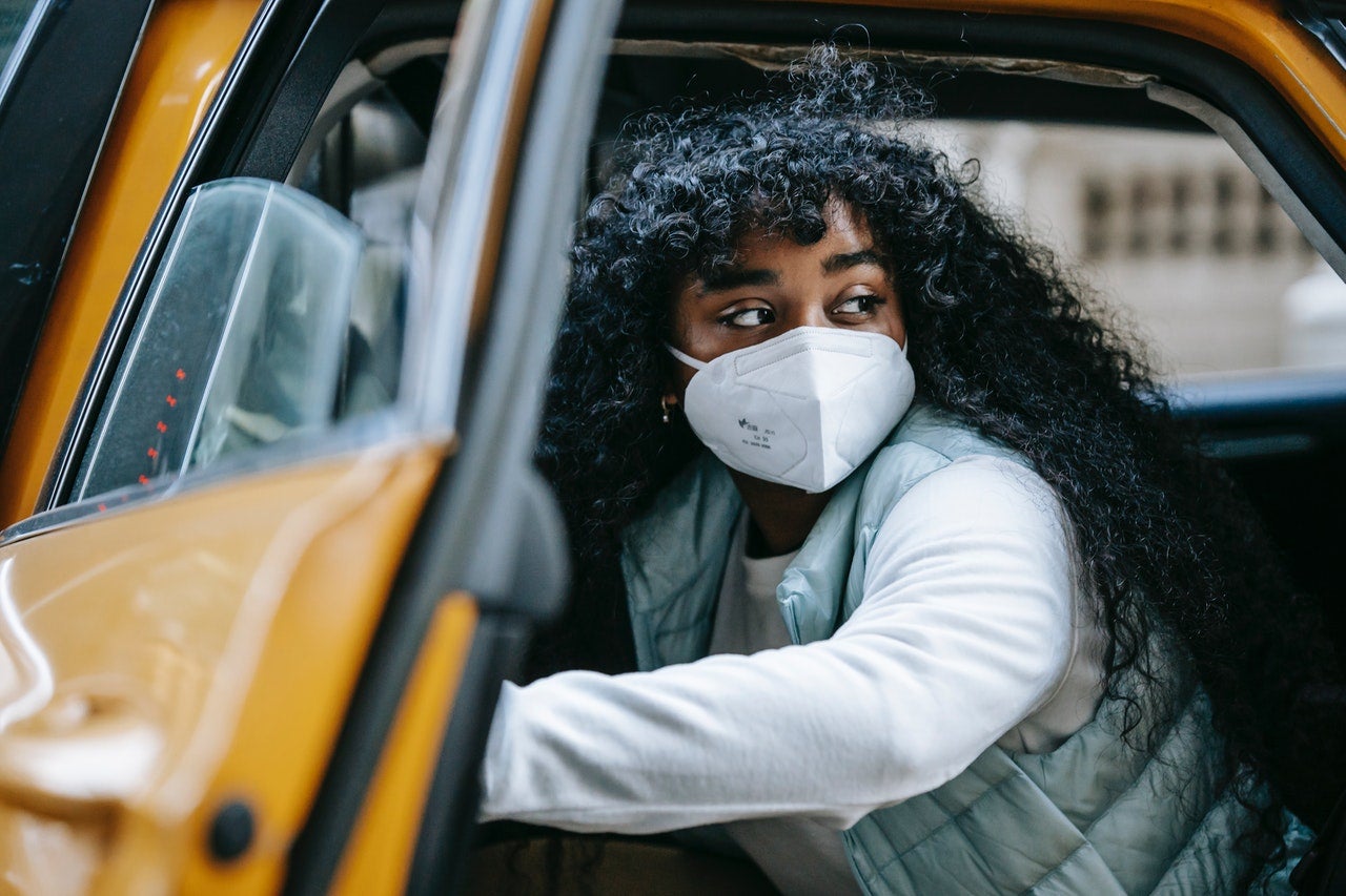 A young Black woman wearing a face mask is stepping out of a yellow Taxi cab and looking over her shoulder.