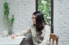 A young Asian woman sits at a tabletop writing in a notebook about how to budget for insurance.