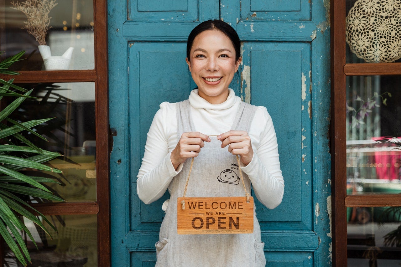 A woman stands in front of her business with a "we're open" store sign.