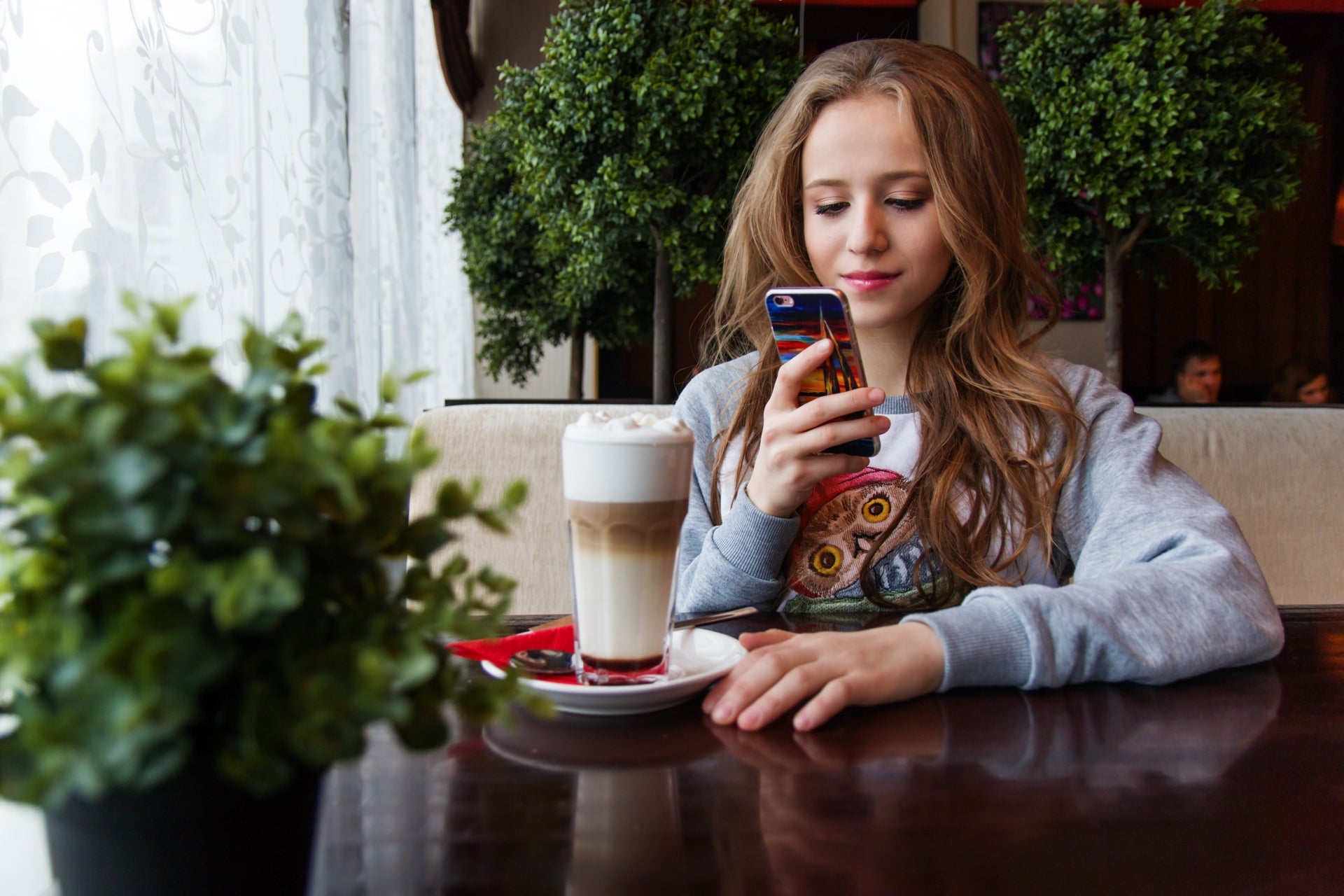 A young woman in a restaurant looks at her cell phone. She is sitting at a table with a milkshake in front of her, and there is greenery around her.
