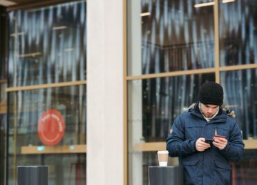 A young man wearing a heavy winter coat and a beanie stands outside a building looking down at his cellphone and holding a credit card.
