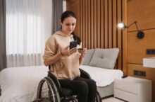 A young woman in a wheelchair next to her bed looks at her phone and holds a credit card.