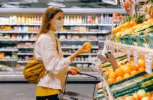 A young white woman wearing a white jacket and a face mask holds a lemon up in a grocery store.
