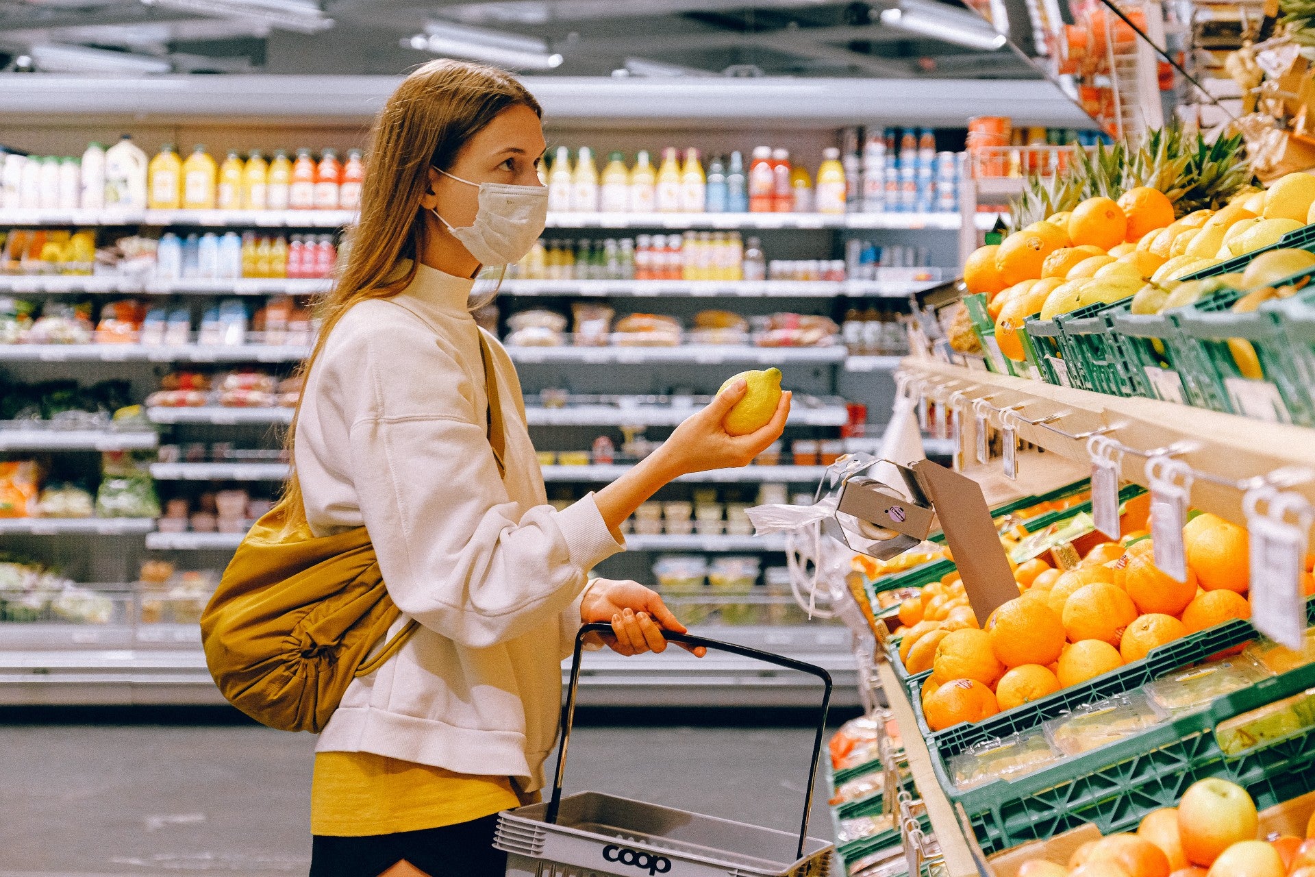 A young white woman wearing a white jacket and a face mask holds a lemon up in a grocery store.