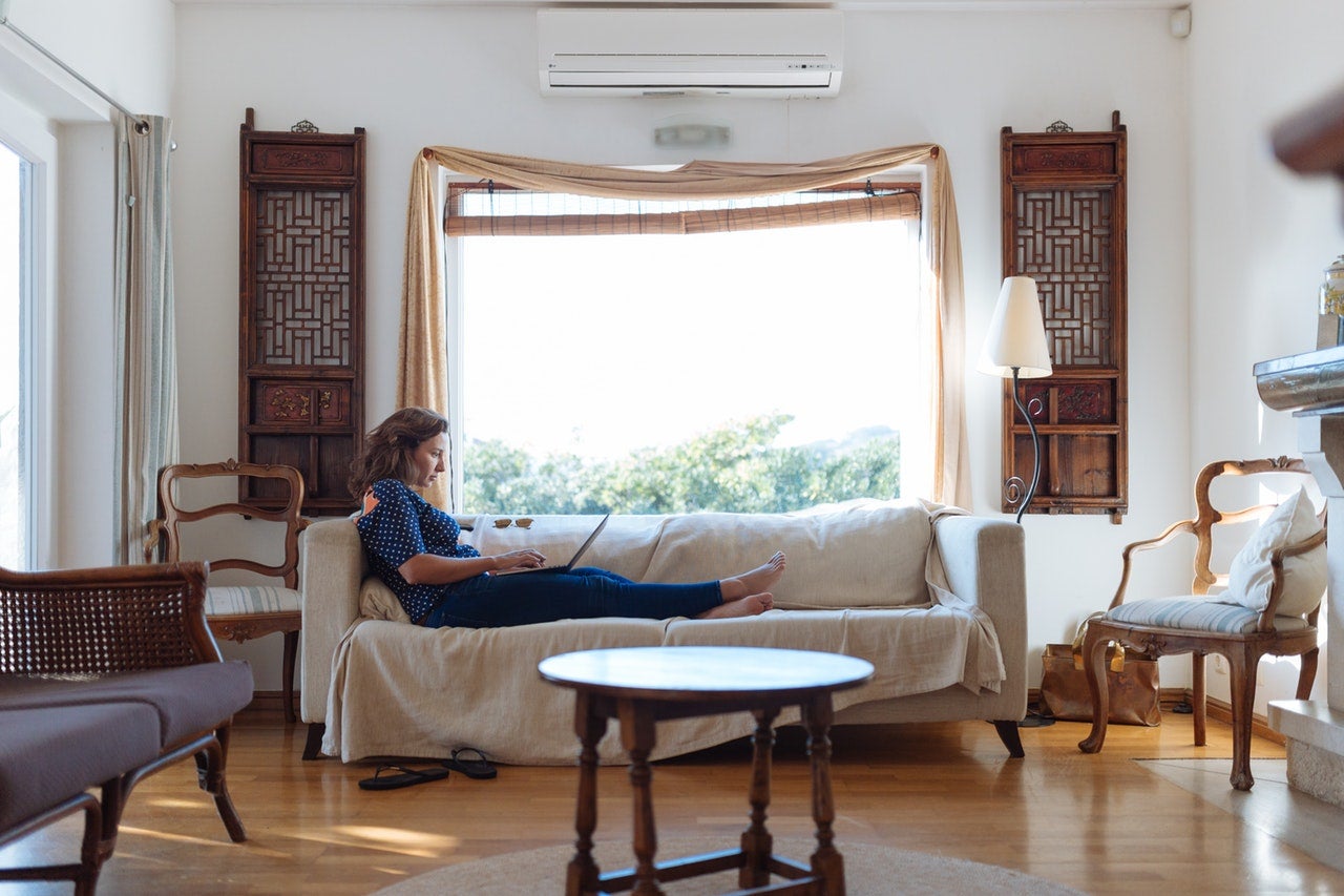 A woman in a gray shirt lounges on the couch in her rented apartment, checking her credit score on her laptop.