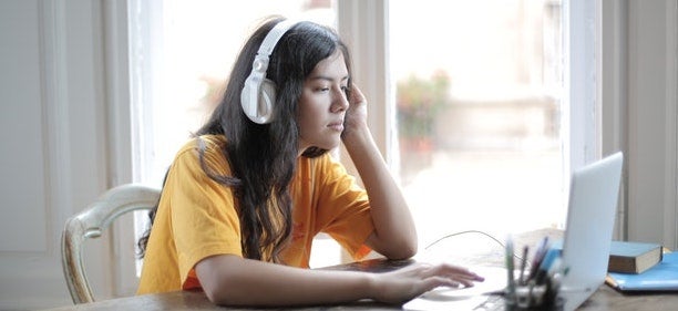 A woman in a yellow shirt wears white headphones as she looks on her laptop.