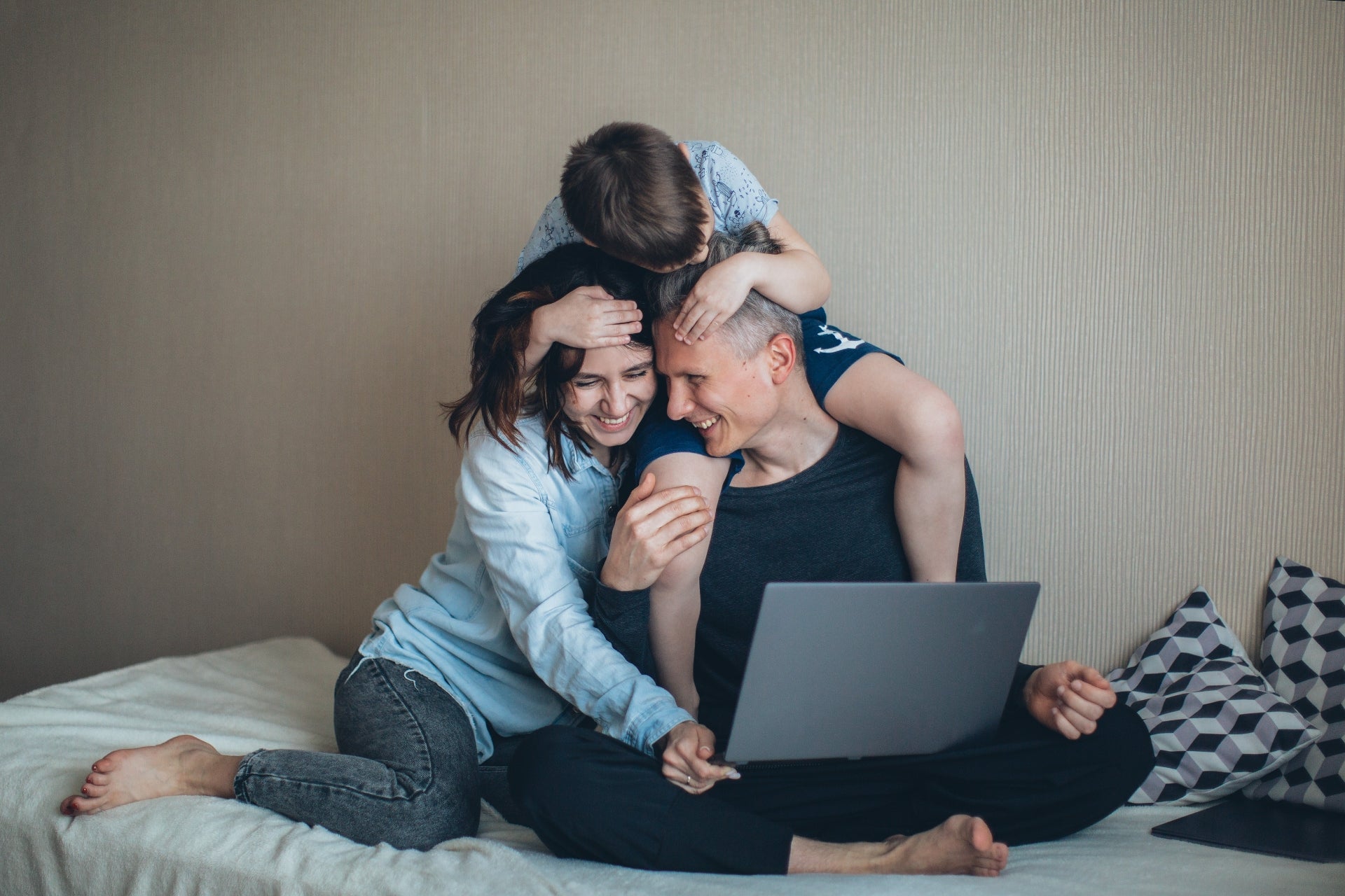 A couple sits on a bed looking at a laptop. Their young son sits on the father's shoulders embracing both of them.