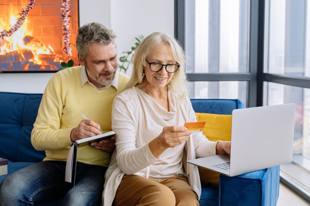 An older white couple sits on a dark blue couch in front of a laptop, smiling and looking at a credit card.