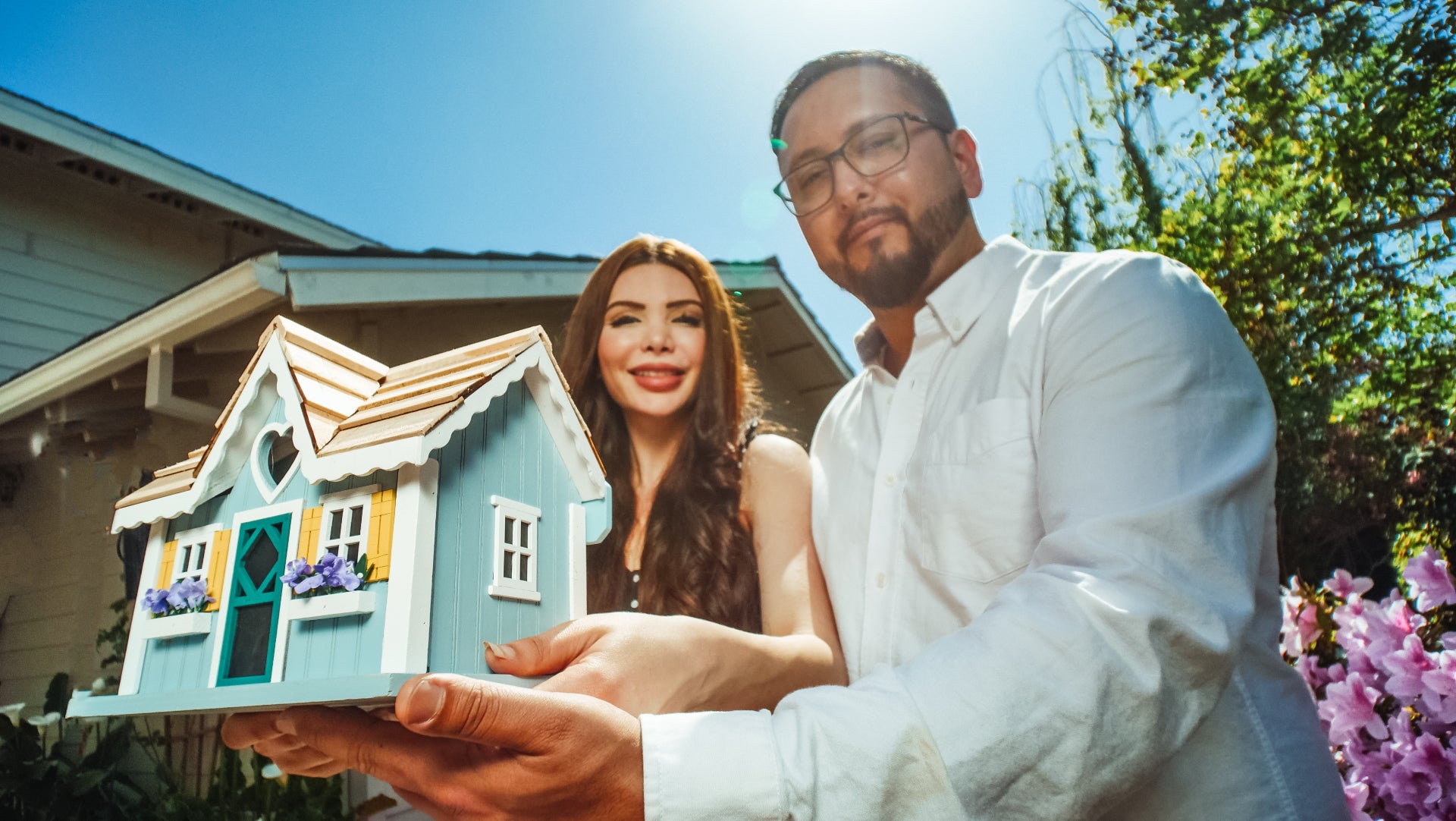 A couple sits in front of their new home, holding a model of a smaller home in front of them