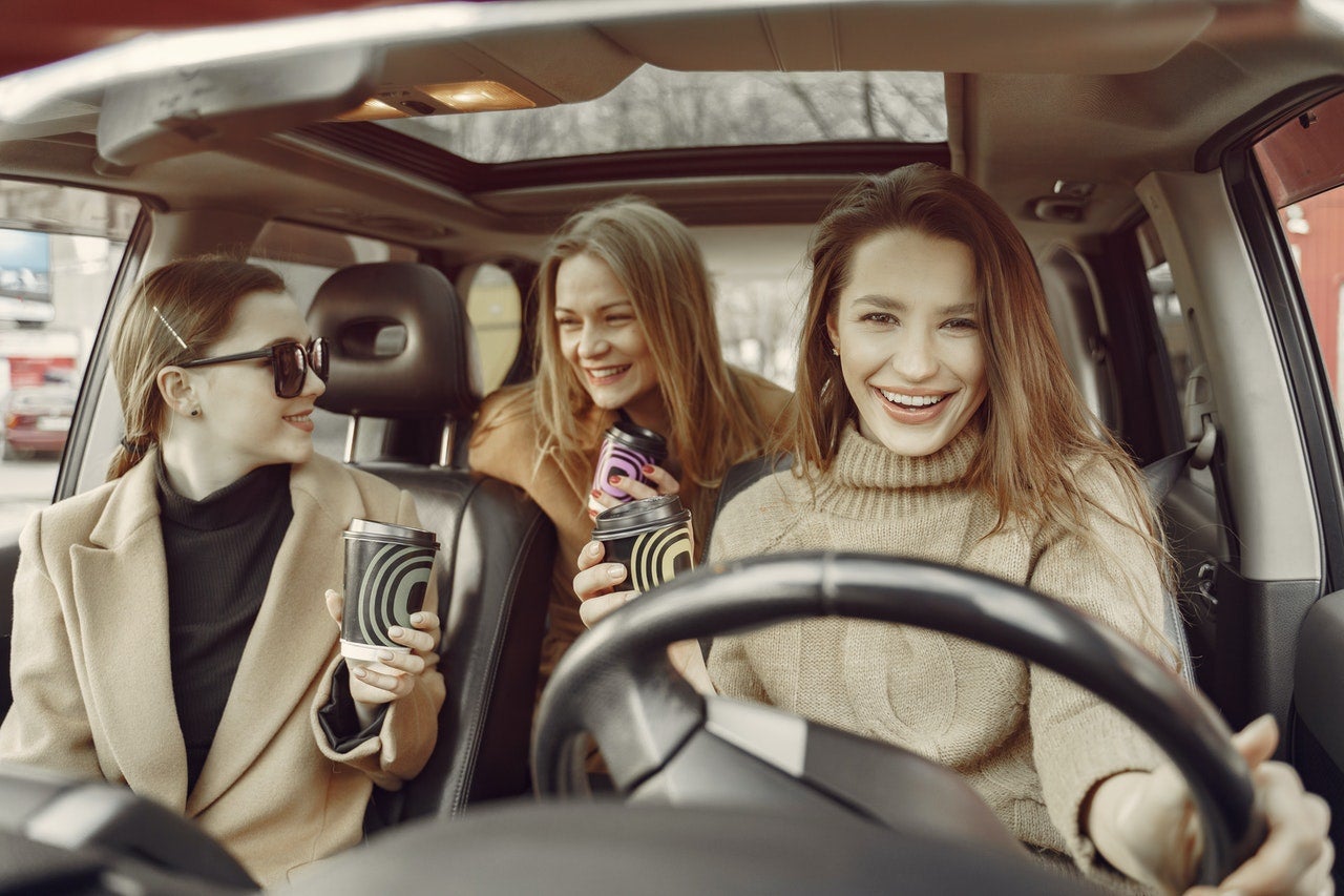 Three friends drive in a car, laughing and drinking coffee