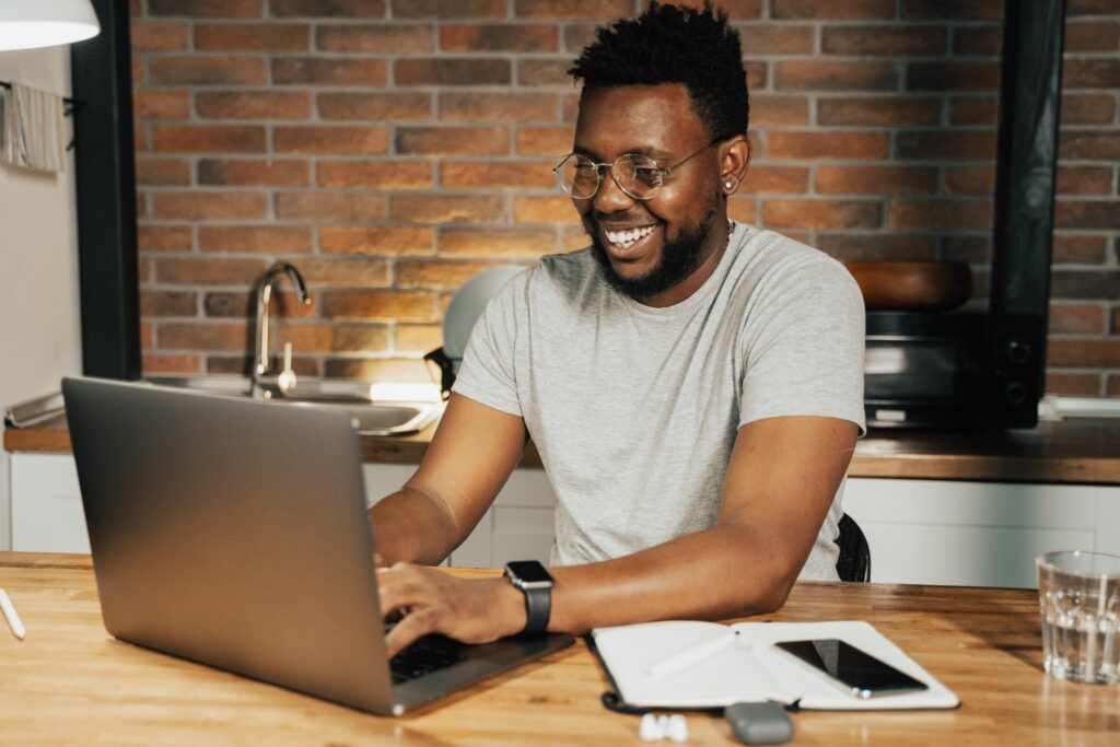 A young Black man with glasses and a gray tshirt sits at his kitchen counter smiling at his laptop learning how to maintain a 700 credit score with late payments.