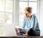 A full length photo of woman using laptop while holding documents at home. Young female is sitting on window sill. She is working from home.