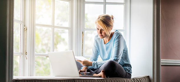 A full length photo of woman using laptop while holding documents at home. Young female is sitting on window sill. She is working from home.