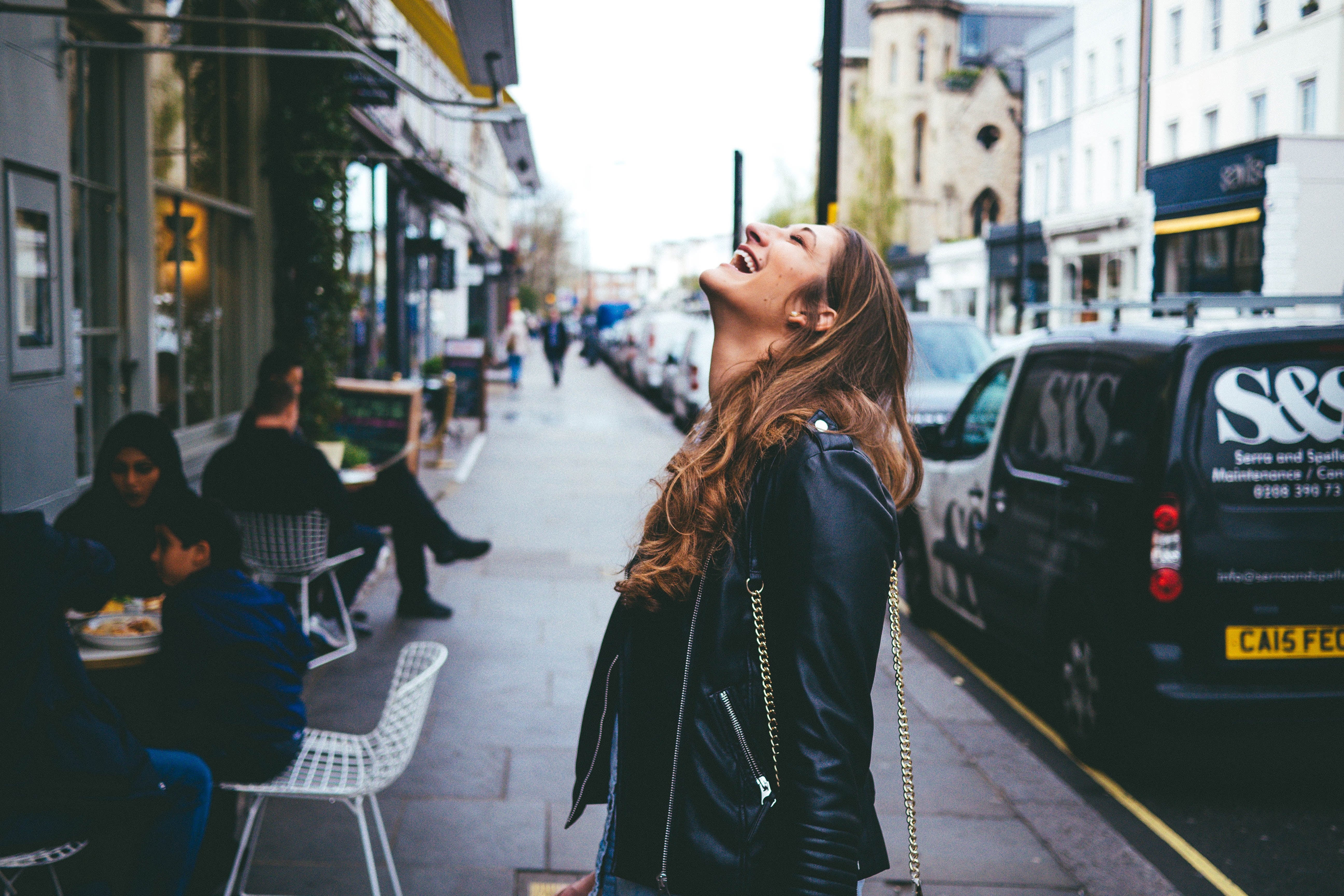 A woman closing her eyes and looking up laughs on the sidewalk outside a cafe.