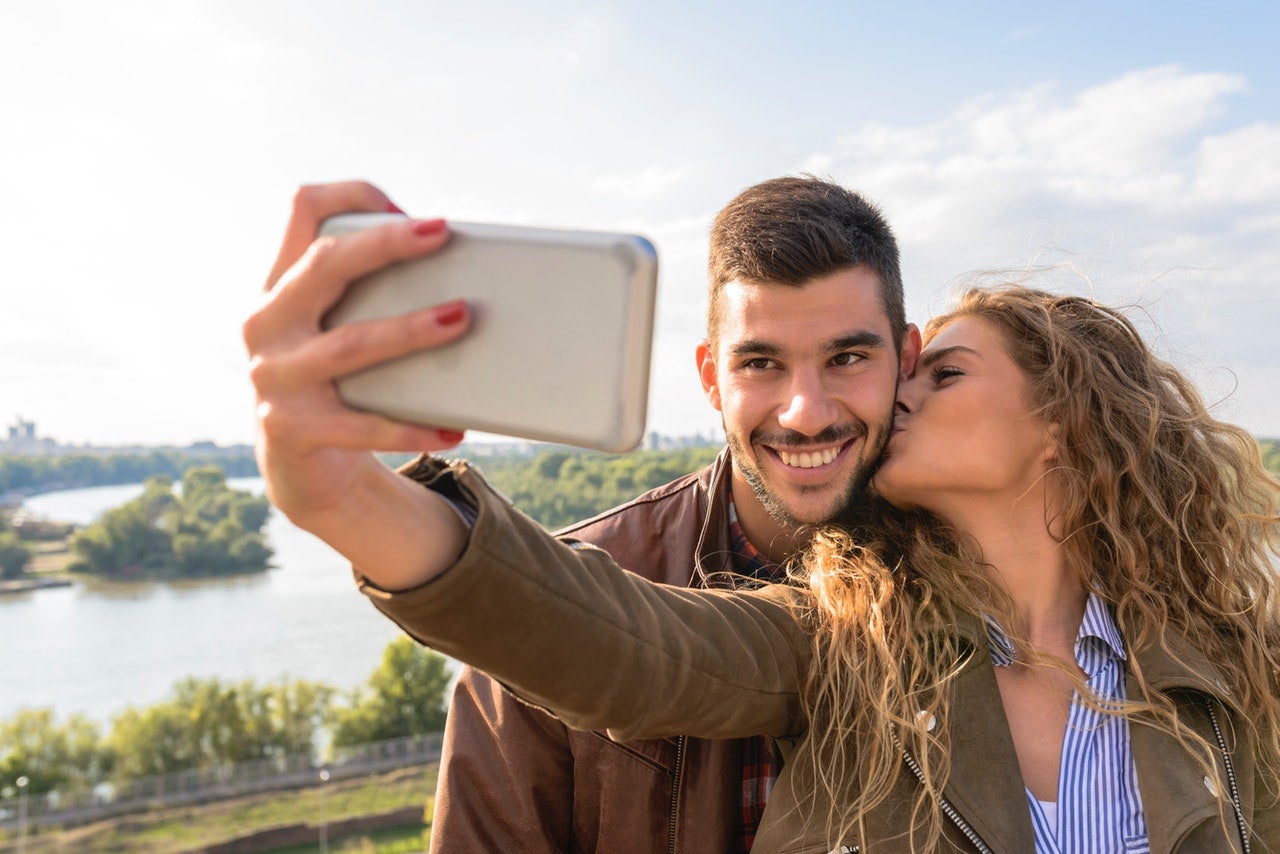 A woman kisses the cheek of a smiling man while taking a selfie of the two of them.