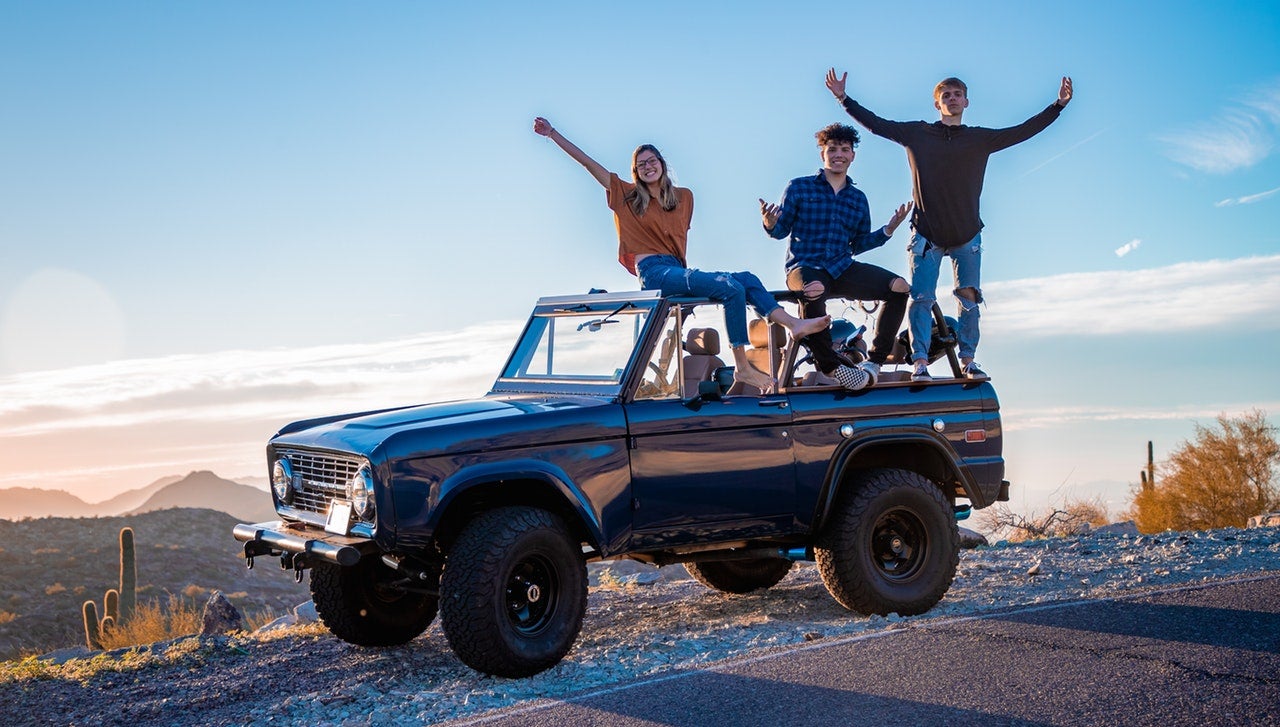 Three young people sit on top of a large offroad vehicle with their arms outstretched.
