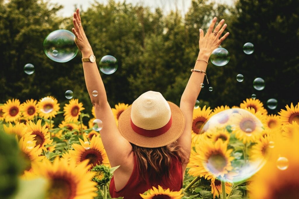A woman in a straw hat stands in a field of sunflowers and bubbles with her back to the camera and her arms raised.