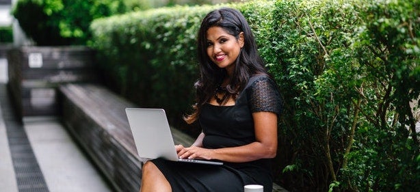 A smiling woman sits on a bench outside with her laptop.