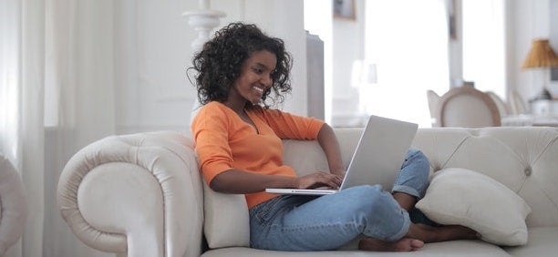 A woman sits on her grey sofa with her laptop on her lap.
