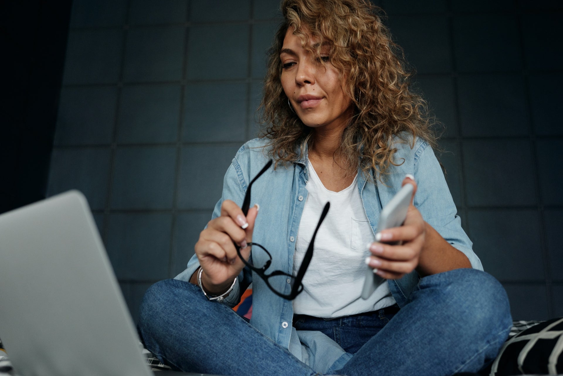 A woman sits crosslegged in front of a computer with her glasses and phone in her hands
