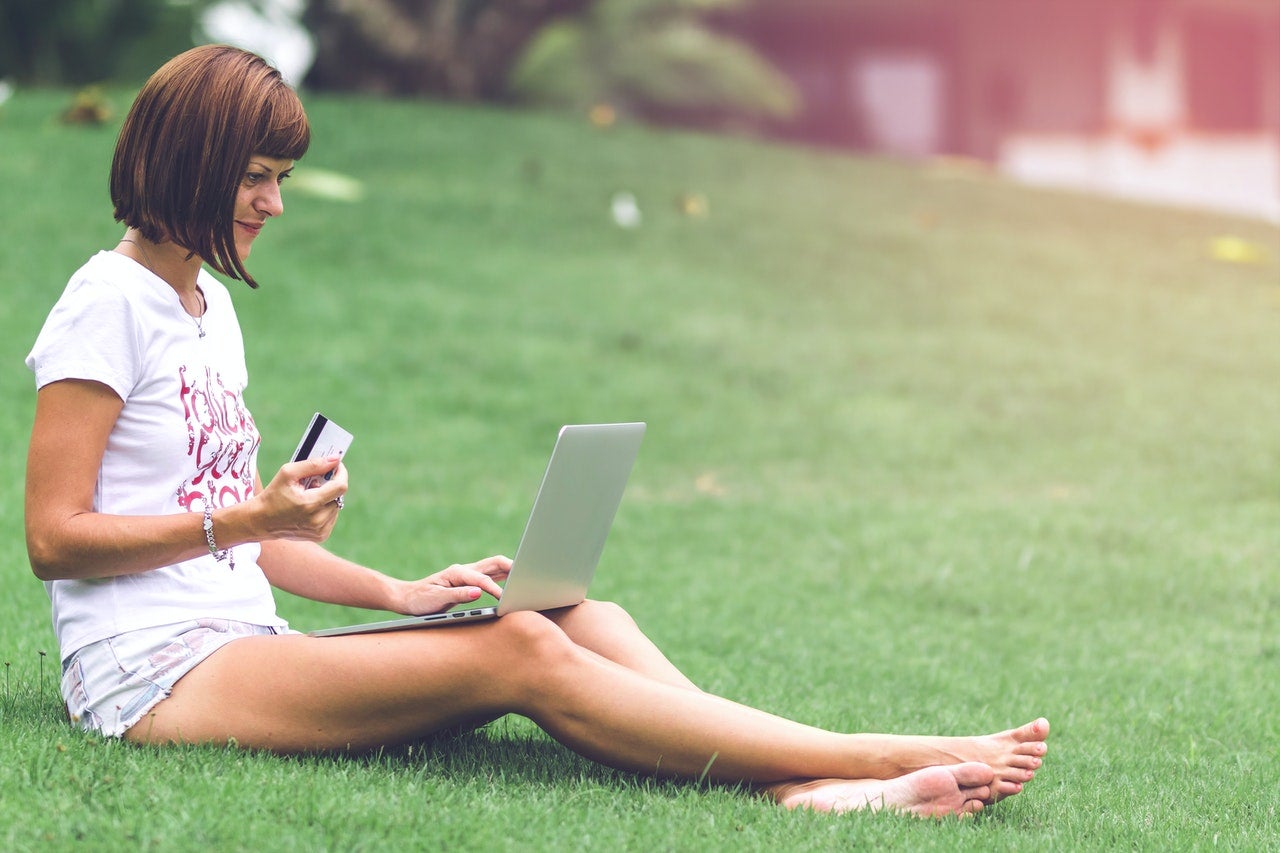 A woman sits on a grassy hill with a laptop on her lap and an OpenSky credit card in her hand.