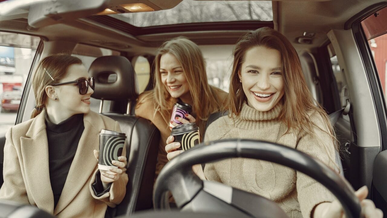 Three friends drive in a car, laughing and drinking coffee