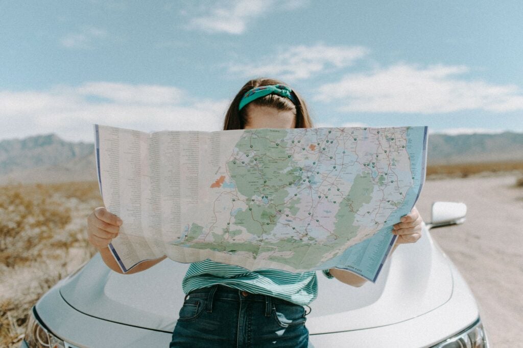 A person sits on the hood of a white car looking at a map. you can only see the very top of their head and their arms holding the map.